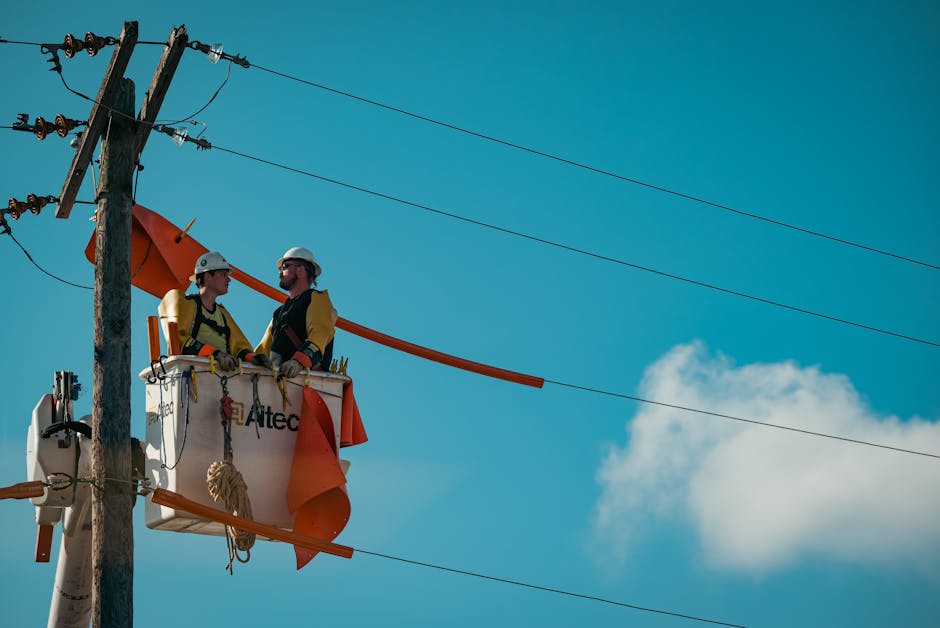 Two electricians in safety gear working on a utility pole with a clear blue sky backdrop.